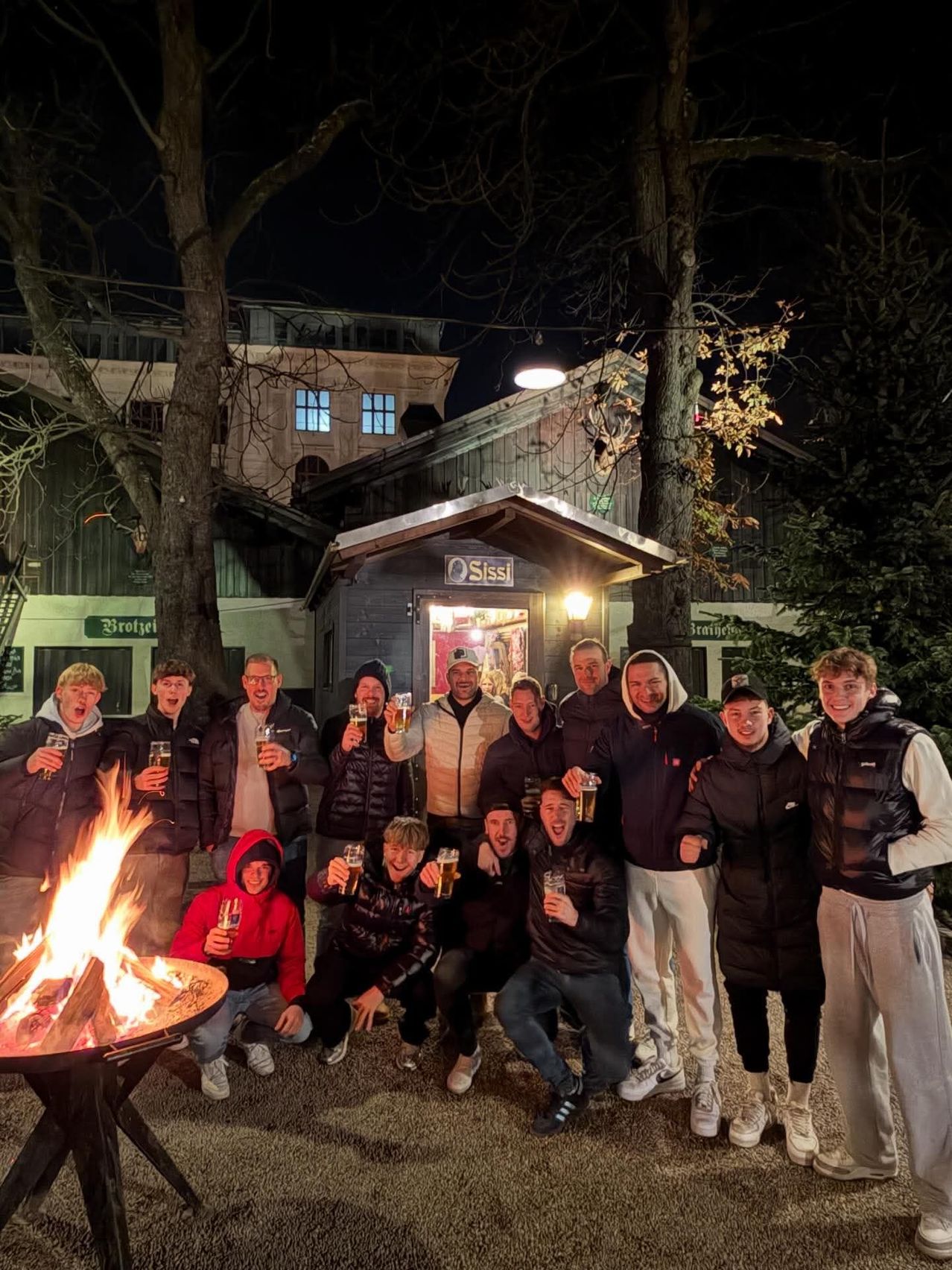 Eine Gruppe von Menschen in Winterkleidung steht um ein Lagerfeuer herum und hält Gläser mit Bier in der Hand. Sie lächeln und scheinen sich zu amüsieren. Hinter ihnen befindet sich ein Gebäude mit einem Schild, auf dem 'Sissi' steht.