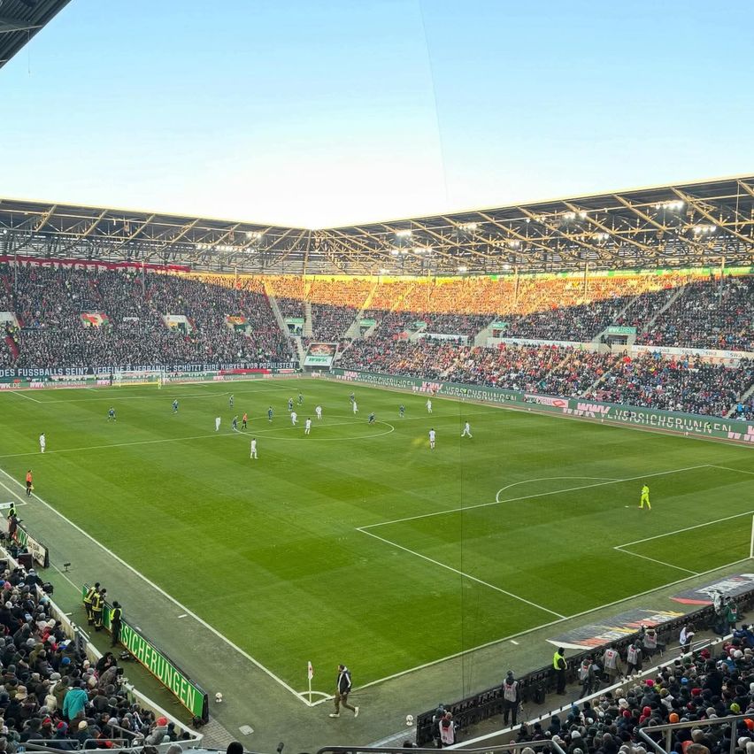 Ein mit Zuschauern gefülltes Fußballstadion bei einem Spiel an einem sonnigen Tag, mit Spielern über das Feld verteilt und einer Bannerseite.