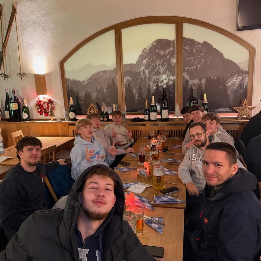 Eine Gruppe junger Männer in einem Restaurant lächelt und posiert für ein Foto. Sie sitzen um einen Tisch mit Flaschen und Gläsern herum. Der Tisch ist mit einem weiß-blau karierten Tuch bedeckt. Hinter ihnen befindet sich ein großes Fenster mit Bergblick.