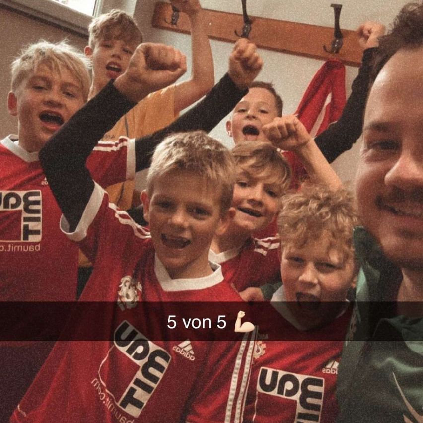 Eine Gruppe von Jungen in roten Trikots mit dem Wort 'Upe' darauf, heben ihre Fäuste und lächeln. Das Bild hat einen Wasserzeichen '5 von 5'.