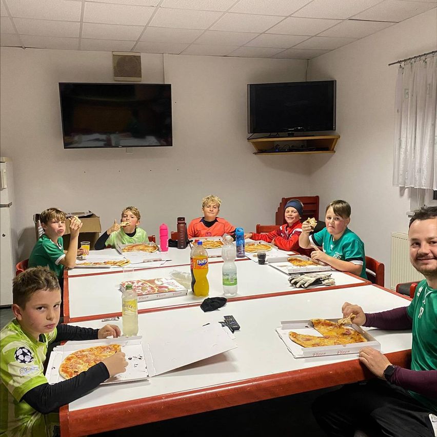 Eine Gruppe junger Jungen isst gemeinsam Pizza in einem Raum mit weißen Wänden, einem Fernseher und einem Kühlschrank.