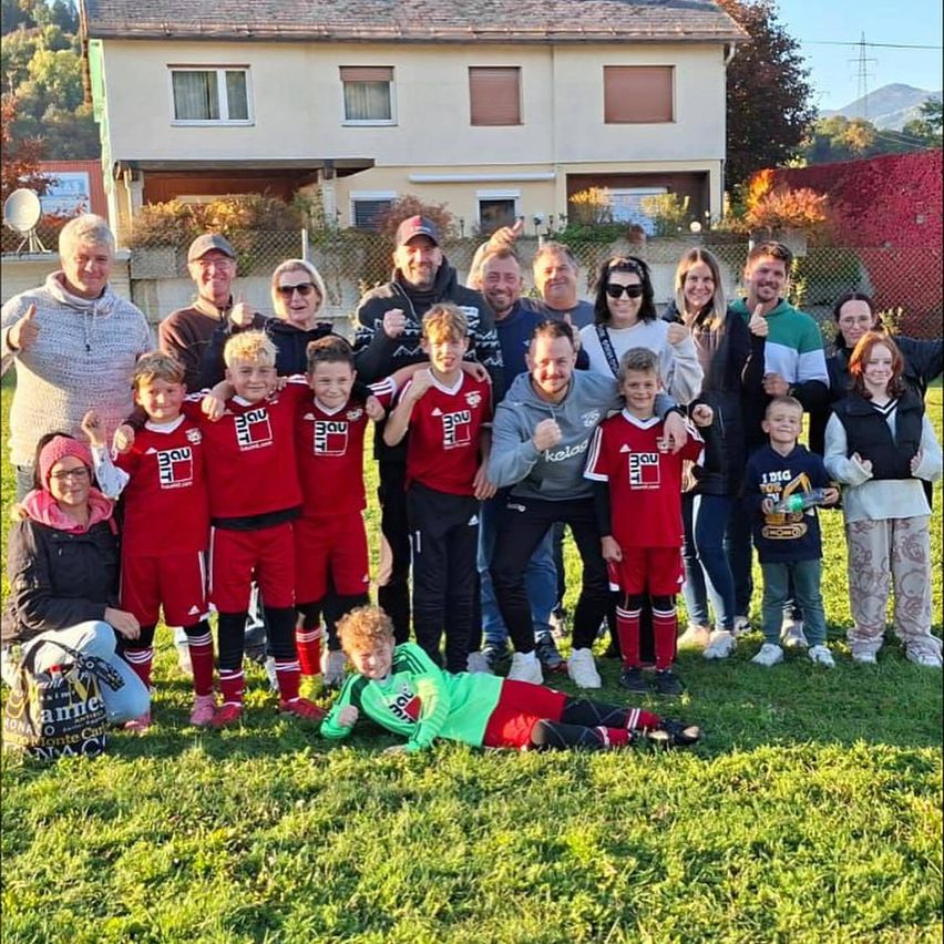 Eine Fußballmannschaft in roten Trikots posiert für ein Foto auf einem Rasenfeld, mit Erwachsenen und Kindern dahinter. Im Hintergrund befindet sich ein Haus mit Pflanzen und einem Zaun.