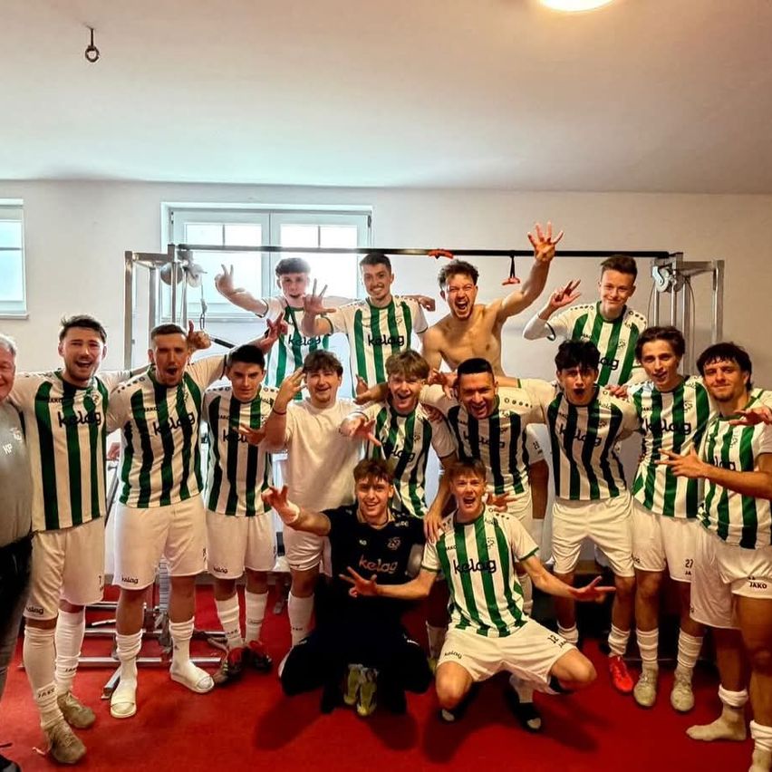A soccer team in green and white uniforms celebrates together in a gym, posing for a photo with a red carpet floor and white walls.