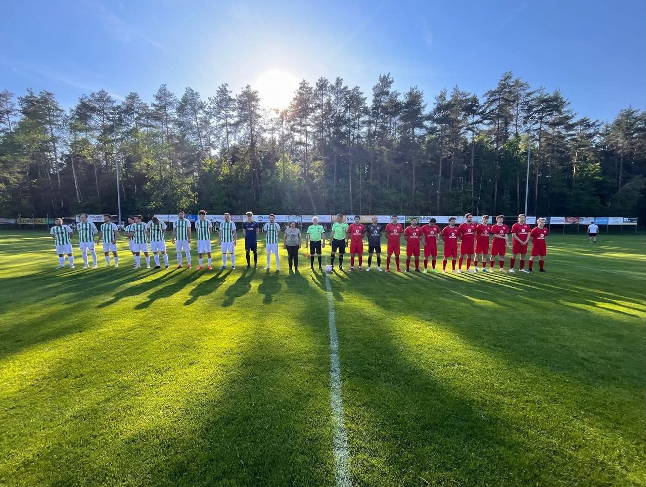 Eine Gruppe von Fußballspielern in grünen und roten Uniformen steht auf einem Feld mit einer weißen Linie, umgeben von Bäumen.