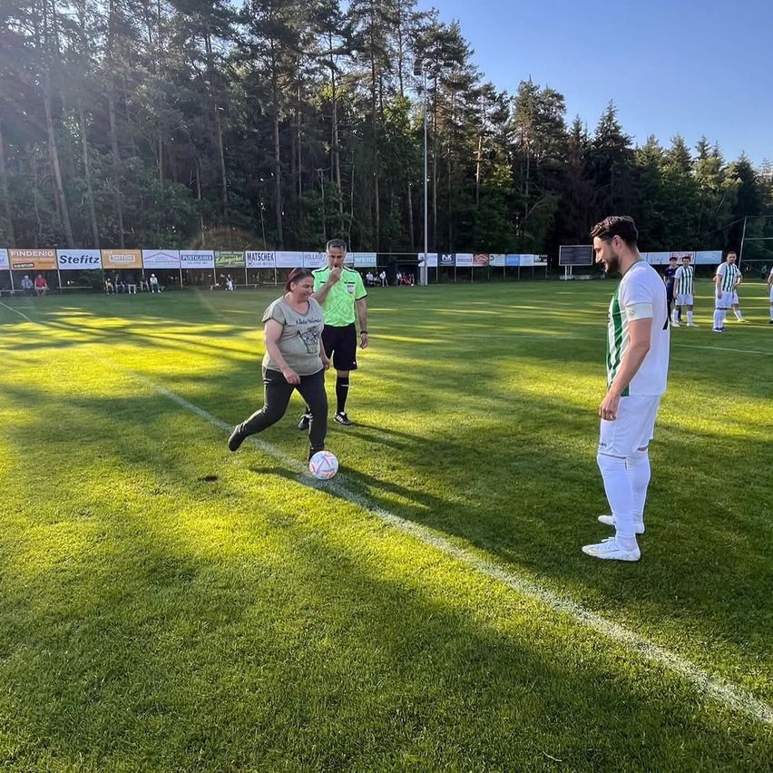 Eine Frau tritt einen Fußball auf einem grünen Feld, während ein Mann und ein Schiedsrichter zusehen. Im Hintergrund befinden sich Spieler und Zuschauer.
