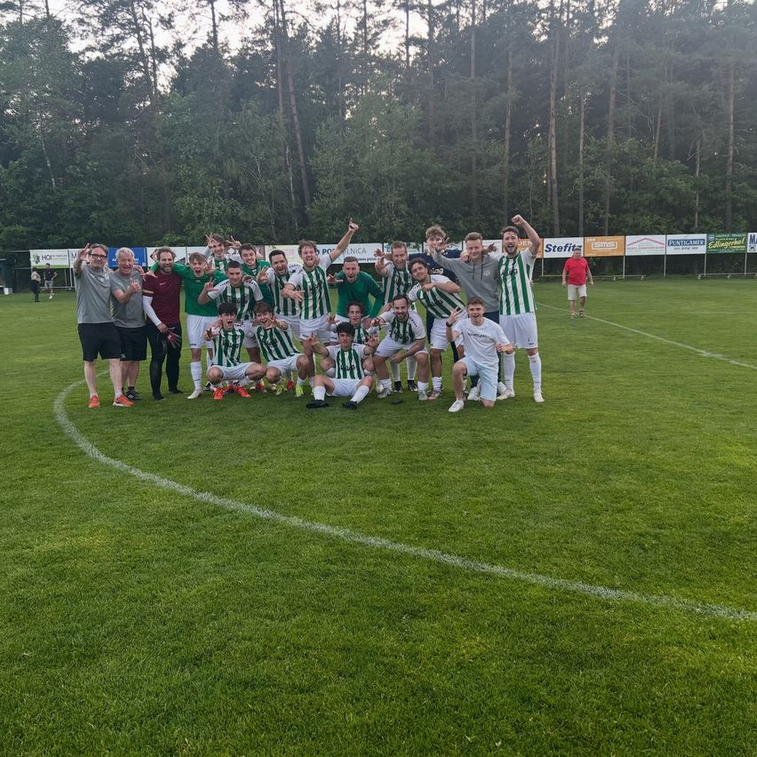Eine Fußballmannschaft posiert auf dem Feld, umgeben von einem üppigen grünen Wald. Spieler in grünen und weißen Uniformen feiern gemeinsam.