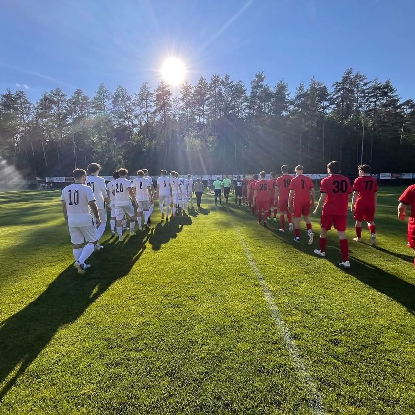 Fußballspieler in weißen und roten Trikots gehen auf das Spielfeld zu, mit einer strahlenden Sonne am Himmel und einer Reihe von Bäumen im Hintergrund.