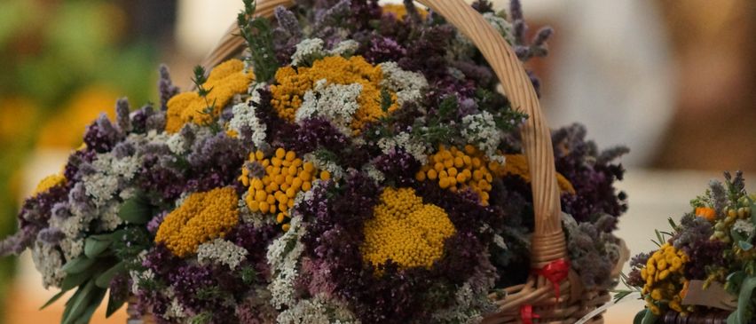 Ein Korb aus Weide gefüllt mit einer Mischung aus lila und gelben Blumen steht auf einem Marmoraltar. Im Hintergrund ist eine verschwommene Figur zu sehen, und eine Kerze ist angezündet.