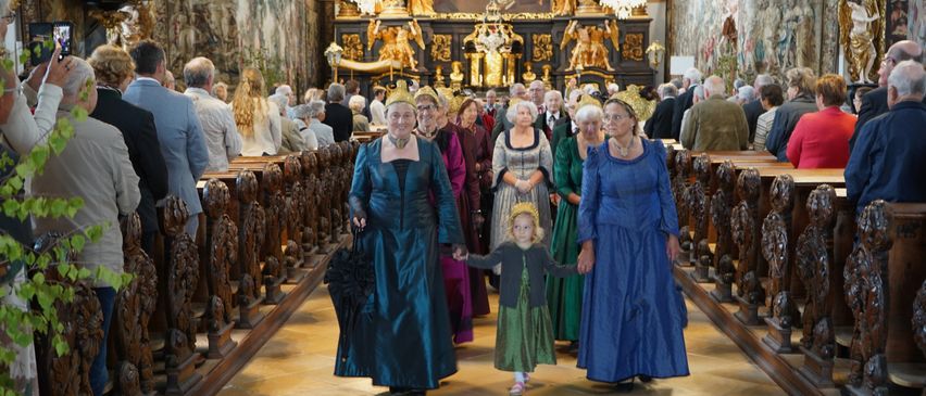 Eine Gruppe von Frauen und einem Kind in historischer Kleidung geht durch den Gang einer Kirche, mit einem kunstvollen Altar und Kronleuchtern im Hintergrund.
