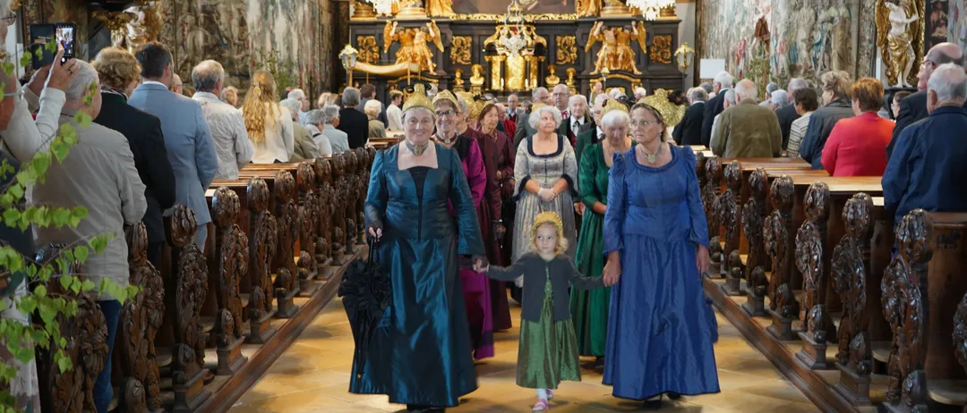 Eine Gruppe von Frauen und einem Kind in historischer Kleidung geht durch den Gang einer Kirche, mit einem kunstvollen Altar und Kronleuchtern im Hintergrund.