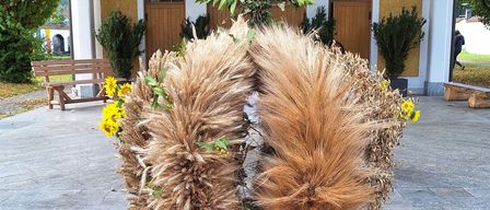 Ein Holzständer mit einer dekorativen Anordnung aus getrockneten Weizenähren, gelben Blumen und kleinen Kürbissen. Der Ständer steht auf einem Kopfsteinpflaster vor einem Gebäude.