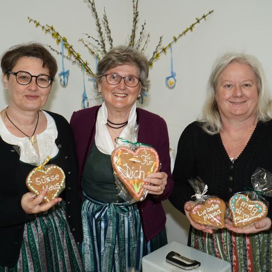 Drei Frauen halten herzförmige Kekse, einer mit 'Süsse Maus', einer mit 'Für Dich' und einer mit 'Liebe Mama'. Sie lächeln und stehen vor einer weißen Wand mit dekorativen Zweigen.