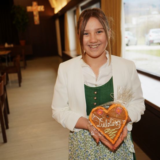 Ein junges Mädchen in traditioneller Kleidung hält einen herzförmigen Lebkuchen mit dem Wort 'Liebling' darauf. Sie lächelt und steht in einem Raum mit Holzstühlen und einem Kreuz an der Wand.