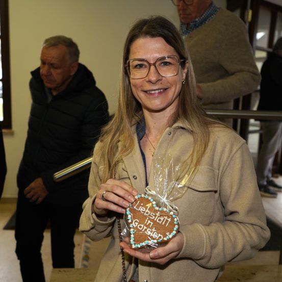 Eine Frau mit Brille hält einen herzförmigen Keks mit den Worten 'Liebsteiditt' und 'Garten' darauf. Hinter ihr stehen zwei Männer, einer von ihnen trägt einen braunen Pullover.