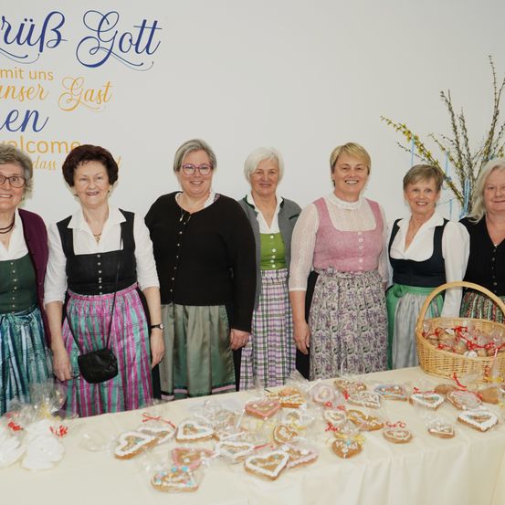 Eine Gruppe von Frauen in traditioneller Kleidung steht hinter einem Tisch voller dekorierter Kekse, mit einem Banner im Hintergrund.