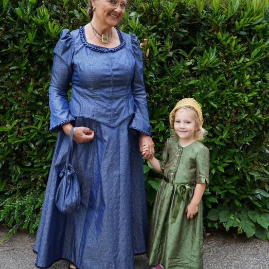 Eine ältere Frau in einem blauen Kleid mit Puffärmeln und einer Halskette steht neben einem kleinen Mädchen in einem grünen Kleid, beide lächeln und halten Händchen vor einem grünen Busch.