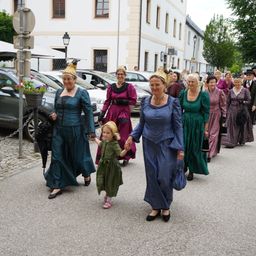 Eine Gruppe von Frauen in Vintage-Kleidung, einschließlich eines jungen Mädchens, geht in einer Reihe auf einer Kopfsteinpflasterstraße. Sie tragen Hüte und haben Handtaschen.