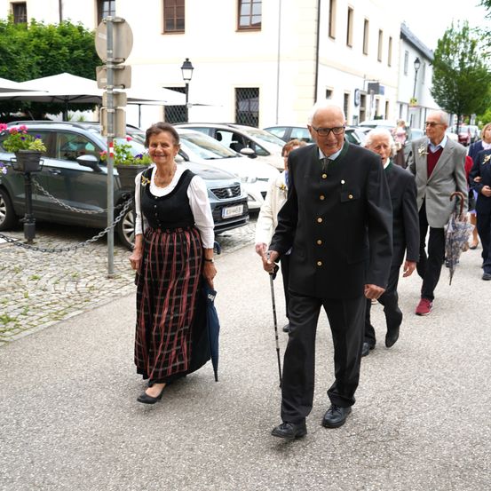Ein älteres Paar in traditioneller Kleidung geht auf einer Kopfsteinpflasterstraße, begleitet von anderen älteren Personen, vor geparkten Autos und Gebäuden.