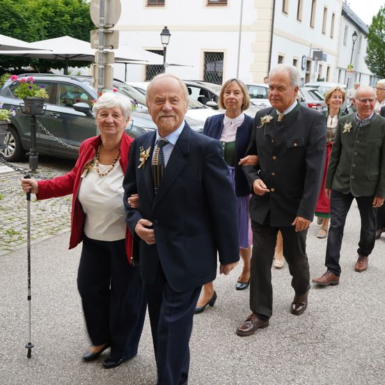 Eine Gruppe älterer Menschen in traditioneller Kleidung geht auf einer Kopfsteinpflasterstraße, möglicherweise bei einem Festival. Sie werden von einer Frau mit einem Stock begleitet. Hinter ihnen sind Autos in der Nähe eines Gebäudes geparkt.