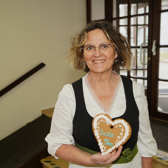 Eine Frau mit Brille und traditioneller Kleidung hält einen herzförmigen Keks mit der Aufschrift 'Beste Oma'.