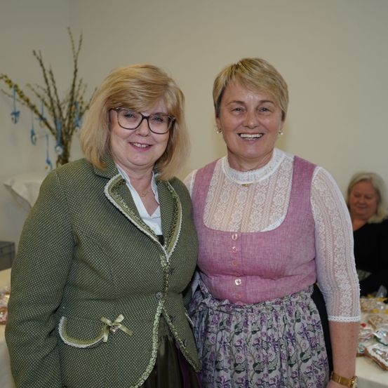 Zwei Frauen lächeln und stehen nebeneinander, eine trägt eine grüne Jacke und die andere ein rosa Oberteil mit einem Blumenrock. Hinter ihnen steht ein Tisch mit verschiedenen Gegenständen und eine Frau im Hintergrund.