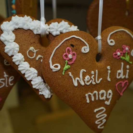 Drei herzförmige Lebkuchen hängen, jeweils mit dekorativer Glasur. Der mittlere hat eine rosa Blume und die Worte 'Weil i di mag' darauf geschrieben. Der Cookie auf der linken Seite hat weiße Glasur und das Wort 'ste'. Der Cookie auf der rechten Seite hat rosa Blumen und ein Herz.
