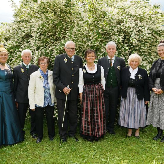 Eine Gruppe älterer Menschen in traditioneller Kleidung posiert für ein Foto vor einem großen blühenden Busch.