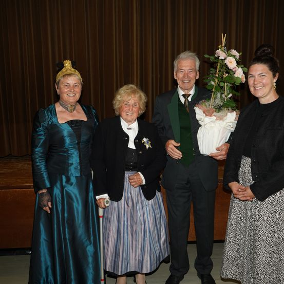 Vier Personen stehen lächelnd zusammen, ein Mann hält einen Blumenstrauß. Eine Frau links trägt ein blaues Kleid und eine Krone, eine andere Frau rechts trägt ein schwarzes Kleid. Der Mann in der Mitte trägt ein grünes Weste und eine Krawatte.