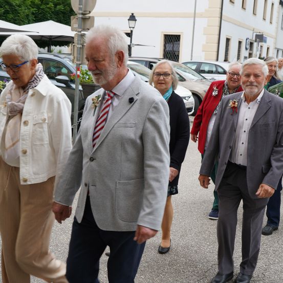 Eine Gruppe älterer Personen, in formeller Kleidung gekleidet, geht eine Straße entlang, gesäumt von geparkten Autos und Gebäuden.