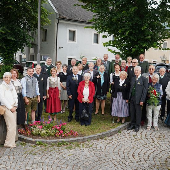 Eine Gruppe älterer Menschen in traditioneller Kleidung posiert für ein Foto vor einem Haus mit Bäumen und einem Blumenbeet.