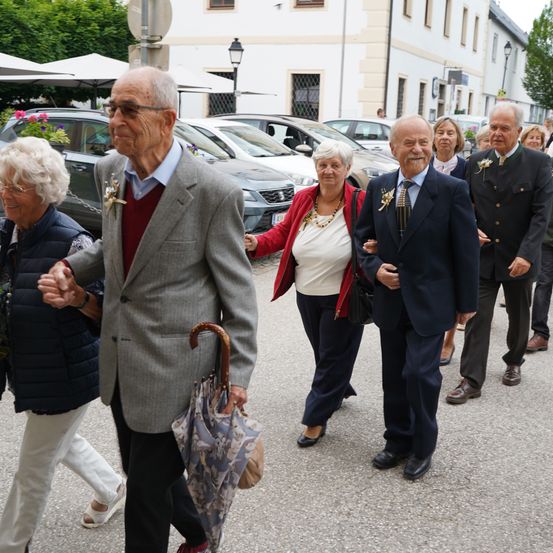 Eine Gruppe älterer Menschen, wahrscheinlich eine Familie, geht eine Straße entlang, mit geparkten Autos dahinter. Sie sind formell gekleidet.