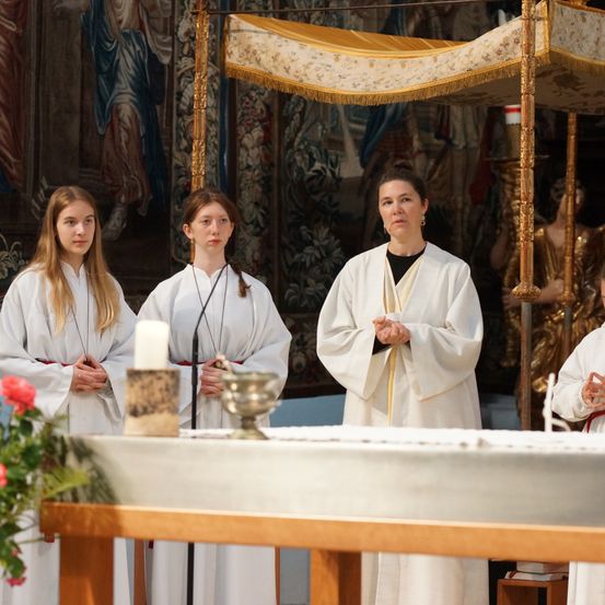 Drei Frauen in weißen Gewändern stehen vor einem Altar, eine hält eine Kerze, mit einem goldenen Baldachin darüber und einem Wandbild im Hintergrund.