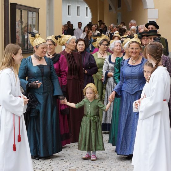 Eine Gruppe von Erwachsenen und Kindern in historischen Kostümen, möglicherweise für ein Fest, gehen in einer Reihe. Die Erwachsenen tragen aufwendige Kopfbedeckungen und die Kinder halten sich an den Händen.