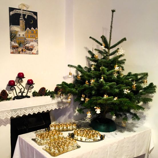 Ein kleiner Weihnachtsbaum mit Engel-Schmuckstücken steht auf einem Tisch mit einer weißen Tischdecke. Auf dem Kaminsims befinden sich zwei rote Kerzen, und ein gerahmtes Bild eines verschneiten Dorfes ist an der Wand. Tabletts mit kleinen Engelfiguren stehen auf dem Tisch.