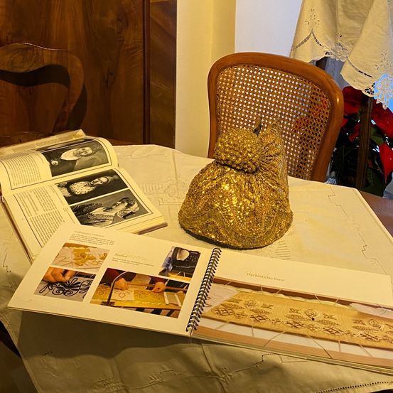 Ein Tisch mit einer goldenen Tasche, einem offenen Buch mit Stickereimustern und einem Stuhl in einem Raum mit einer Holzwand und einer weißen Tischdecke.