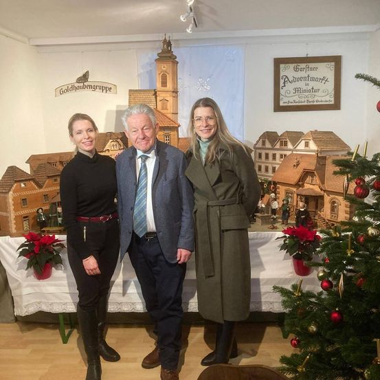 Drei Personen stehen vor einem Weihnachtsbaum und einer Miniaturstadt. Der Mann trägt einen Anzug und eine Krawatte, die Frauen Mäntel und Stiefel. Dahinter steht ein Tisch mit einer roten Weihnachtsblume.