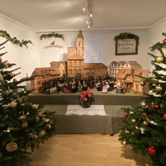 Ein Raum mit einer Weihnachtsdorf-Ausstellung auf einem Tisch, umgeben von zwei geschmückten Weihnachtsbäumen und einem Schild mit der Aufschrift 'Goldhaubegruppe'.