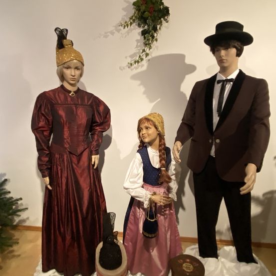 Drei Mannequins in Vintage-Kleidung stehen in einem Raum. Das linke trägt ein burgunderrotes Kleid, das mittlere ein rosa Kleid und das rechte einen braunen Anzug.