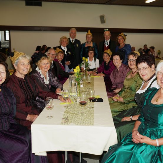 Eine Gruppe älterer Frauen und Männer in Vintage-Kleidung sitzt an einem langen Tisch, lächelt und posiert für ein Foto. Der Tisch ist mit Weingläsern, einer Blumenarrangement und gelben Servietten gedeckt. Der Raum hat weiße Wände und eine Deckenleuchte.
