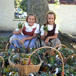 Zwei junge Mädchen in traditioneller Kleidung sitzen auf dem Gras, lächeln und halten Körbe voller Blumen. Sie sind von weiteren Körben mit Blumen umgeben. Dahinter steht ein Baumstamm und ein Gebäude mit einem Fenster.