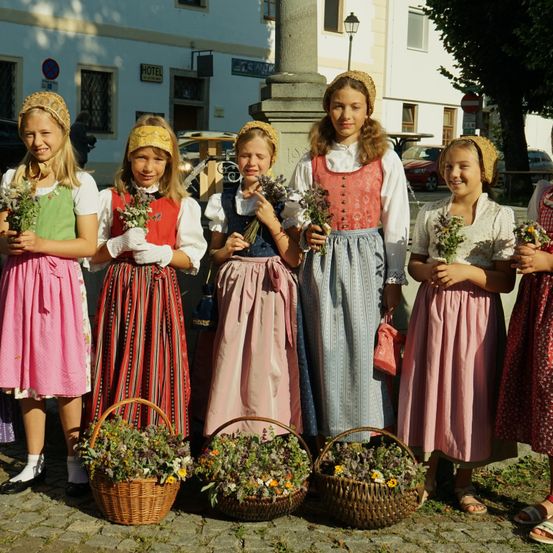 Fünf junge Mädchen in traditionellen Volkstrachten, die Blumenkörbe halten und in einer Reihe stehen. Dahinter befindet sich ein Gebäude mit einem Schild und geparkten Autos.