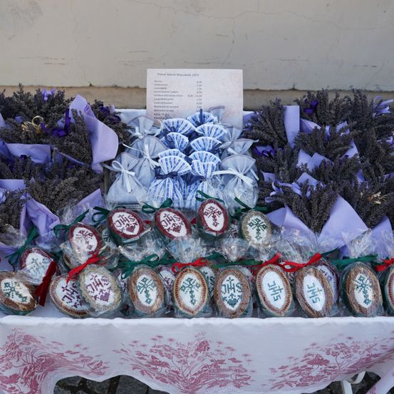 Ein Tisch zeigt eine Auswahl an Lavendel und verpackten Keksen mit religiösen Symbolen. Die Kekse sind in Plastik eingewickelt und mit Bändern gebunden, und im Hintergrund befindet sich eine Preisliste.