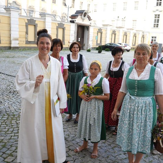 Eine Gruppe von Frauen in traditioneller Kleidung, darunter ein junges Mädchen mit Blumen, geht in einem gepflasterten Innenhof vor einem historischen Gebäude.