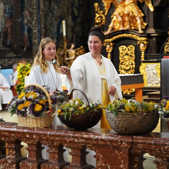 Zwei Frauen in weißen Gewändern stehen in einer Kirche, eine hält ein Mikrofon und die andere einen Korb mit Blumen. Hinter ihnen ist eine goldene Statue und ein Altar mit einer Kerze zu sehen.