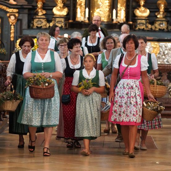 Eine Gruppe von Frauen in traditioneller Kleidung, die Blumenkörbe tragen, geht einen Kirchengang entlang.