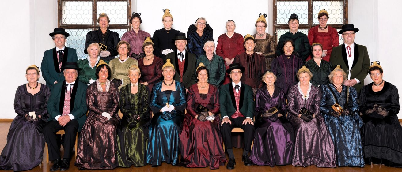 Eine Gruppe von Erwachsenen, die in historischen Kostümen gekleidet sind, wahrscheinlich für eine historische Nachstellung, posieren für ein Foto. Die Kostüme variieren in Farbe und Stil, und einige tragen Hüte.