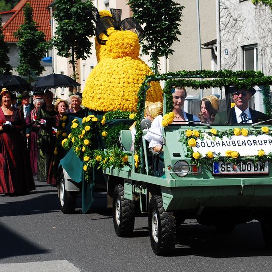 Ein Umzug mit einem großen, blumenbedeckten gelben Fahrzeug, in dem Menschen sitzen, gefolgt von einer Gruppe von Menschen in traditioneller Kleidung mit Regenschirmen.