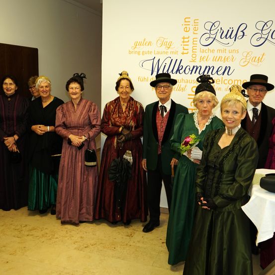 Eine Gruppe von Menschen in Vintage-Kleidung posiert für ein Foto vor einer Wand mit deutschem Text.