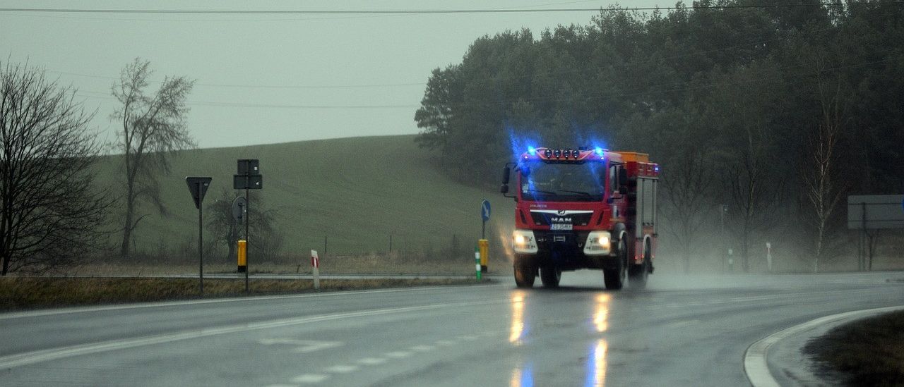 Ein roter Feuerwehrwagen mit blauen Lichtern fährt auf einer nassen Straße, mit Bäumen und Hügeln im Hintergrund.
