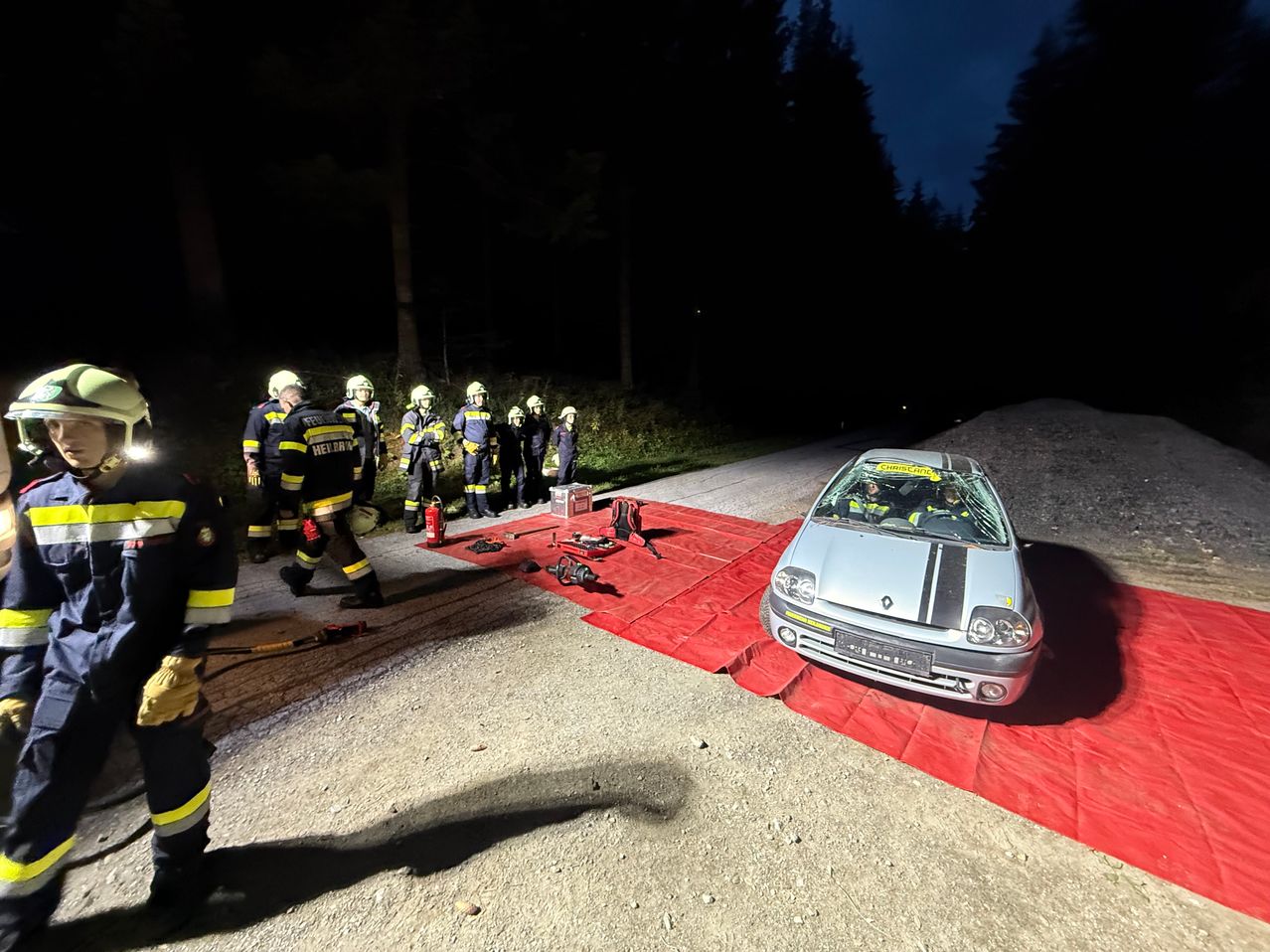 Eine Gruppe Feuerwehrleute in voller Ausrüstung steht um ein beschädigtes Auto auf einer Schotterstraße bei Nacht.